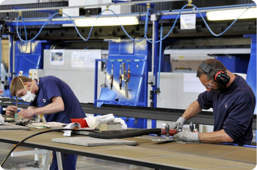 Daher Deux opérateurs Daher travaillent sur une ligne de production. L’un ponce une pièce métallique avec précision, tandis que l’autre prépare un composant sur un établi. Tous deux portent des équipements de protection (masques, gants, casque antibruit) dans un atelier industriel moderne et organisé.